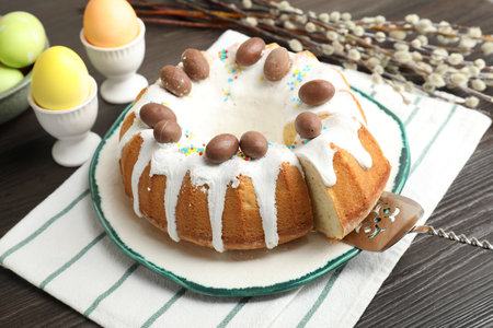 Delicious Easter cake with painted eggs, server and willow branches on wooden table, closeup