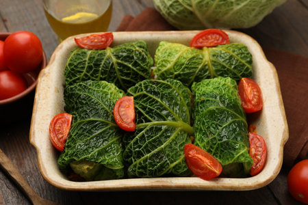 Stuffed cabbage rolls in baking dish and ingredients on wooden table, closeup