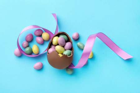 Chocolate Easter egg with candies and ribbon on light blue background, flat lay