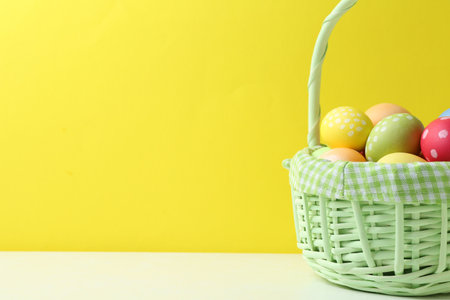 Painted Easter eggs in wicker basket on white table against yellow background, closeup. Space for...