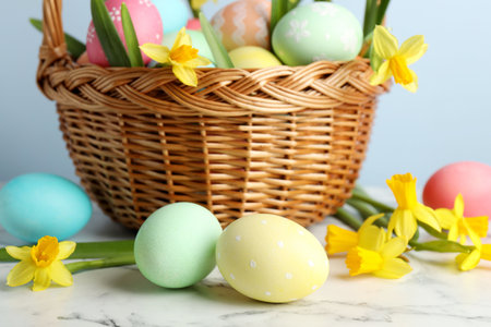 Painted Easter eggs and beautiful daffodils in wicker basket on white marble table against light...