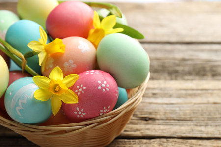 Painted Easter eggs and beautiful daffodils in wicker basket on wooden table, closeup. Space for...