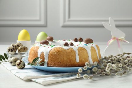 Delicious Easter cake, bunny figure, willow branches, quail and painted eggs on white table indoors,...