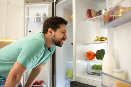 Happy man looking into open refrigerator in kitchenの写真素材