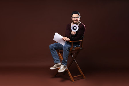 Professional film director with megaphone and script on chair against brown backgroundの写真素材