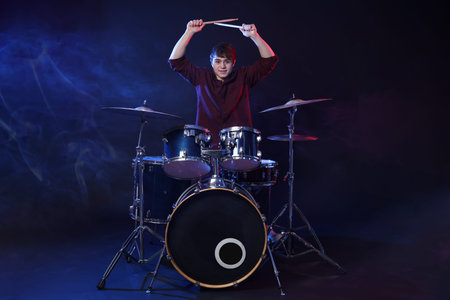 Smiling teenage boy with drumsticks learning how to play drums in color lights against dark backgroundの写真素材