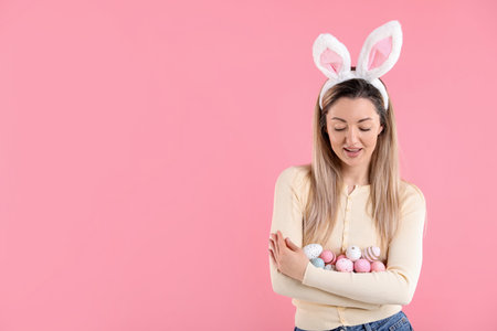 Easter celebration. Smiling woman in headband with bunny ears holding painted eggs on pink...