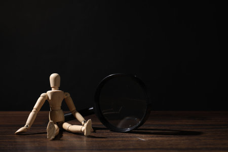 Mannequin with magnifying glass on wooden table against black background, space for textの写真素材