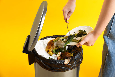 Woman throwing cucumber peels into garbage can on yellow background, closeup