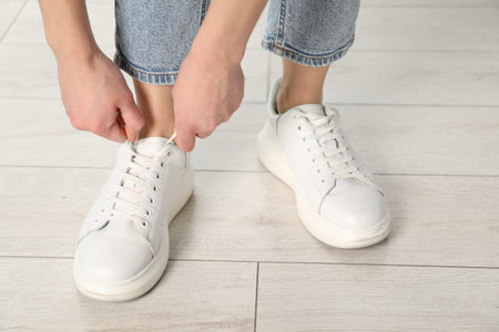 Woman tying shoelace of white sneaker indoors, closeup