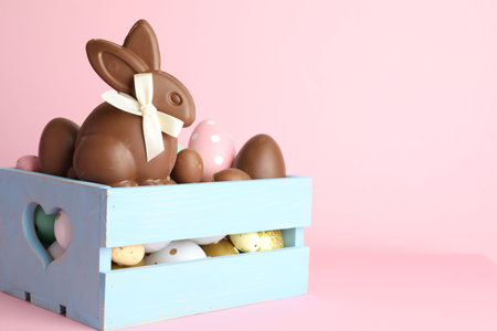 Chocolate Easter bunny and eggs in crate on pink background, closeup. Space for text
