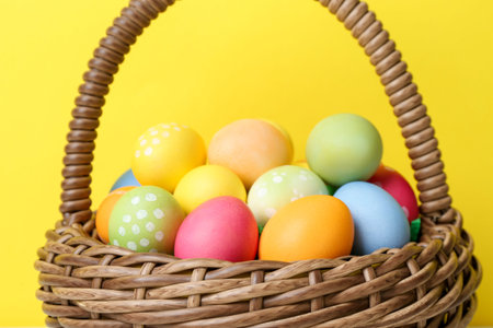 Painted Easter eggs in wicker basket against yellow background, closeup