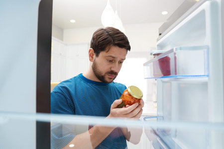Man with jar of pickles near open refrigerator in kitchen, view from insideの写真素材