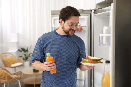 Man with sandwiches and juice near open refrigerator in kitchen