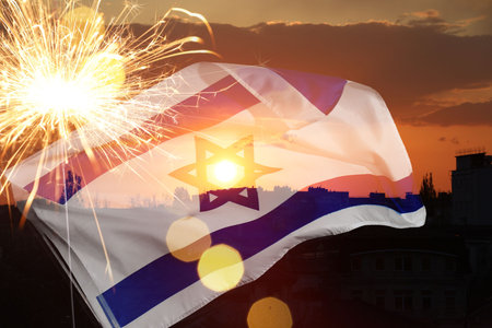 Flag of Israel, burning sparkler and bokeh lights, double exposure with sunset sky above city. National holiday celebrationの写真素材