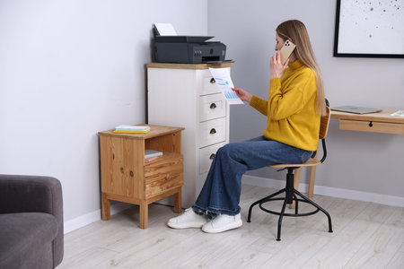Woman talking by smartphone and looking at printout near grey wall indoors