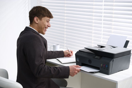 Smiling man taking printed sheet of paper from printer at white desk near window in office