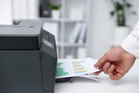 Man taking printed sheet of paper from printer at white desk indoors, closeup