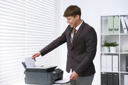 Smiling man working with printer at white desk near window in office