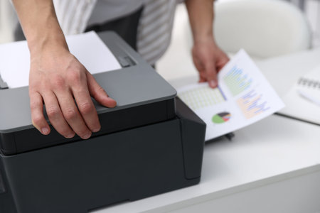 Man taking printed sheet of paper from printer at white desk indoors, closeup