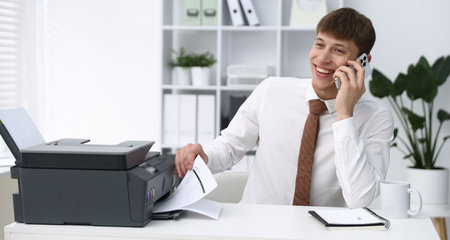 Smiling man taking printed sheet of paper from printer and talking by smartphone at white desk in...