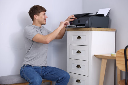 Smiling man taking printed sheet of paper from printer at chest of drawers near grey wall indoors