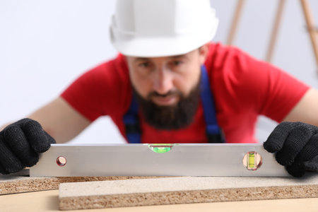 Worker measuring wooden plank with building level at table indoors, selective focus