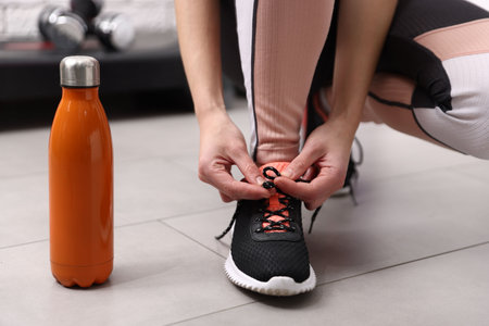 Woman tying shoelace of black sneaker near bottle indoors, closeup