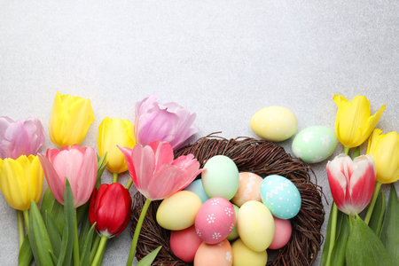 Painted Easter eggs in nest and beautiful tulips on gray textured table, flat lay. Space for text