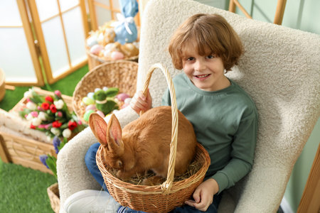 Little boy holding wicker basket with cute Easter bunny in armchair indoors