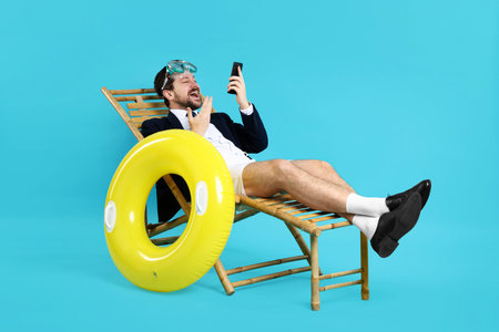 Office worker with diving mask and smartphone in deck chair on light blue backgroundの写真素材