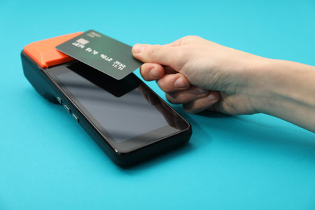 Woman with credit card using payment terminal on light blue background, closeup