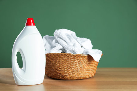 Laundry basket with towels and detergent on wooden table against dark green background, space for...