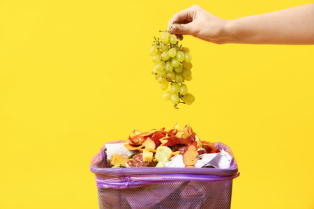 Woman throwing bunch of grapes into garbage can on yellow background, closeupの写真素材
