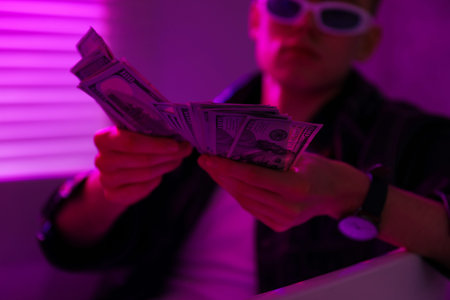 Young businessman in jacket and sunglasses with dollar banknotes in bath tub in violet light...