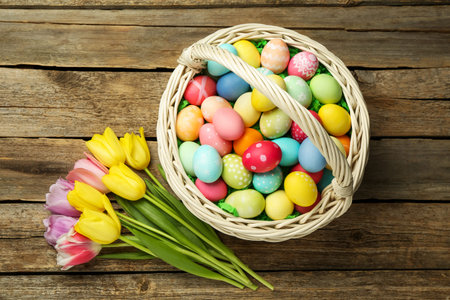 Easter basket with painted eggs and tulips on wooden table, flat lay