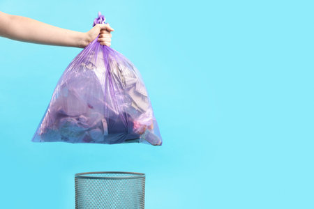 Woman with plastic bag full of garbage and trash bin on light blue background, closeup. Space for...