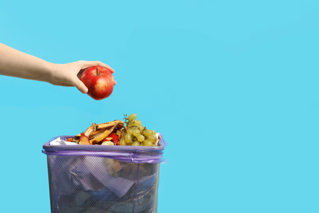 Woman throwing apple into garbage can on light blue background, closeup. Space for text