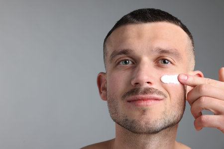 Man applying cream onto his face against light grey background. Space for text