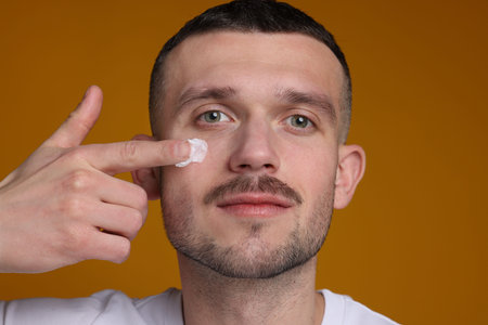 Handsome man applying cream onto his face against orange background