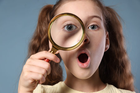 Surprised little girl with magnifying glass on light blue backgroundの写真素材
