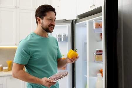 Man with bell pepper and raw meat near open refrigerator in kitchenの写真素材