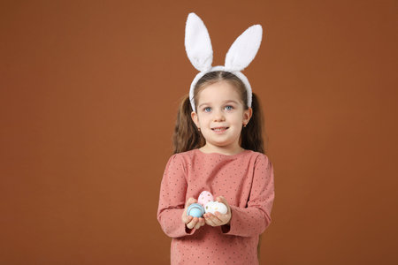 Easter celebration. Smiling little girl in headband with bunny ears holding painted eggs on brown...