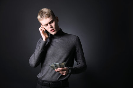Young businessman with dollar banknotes talking by smartphone on dark background
