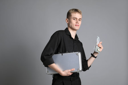 Young businessman in shirt with metal briefcase and dollar banknotes on gray backgroundの写真素材