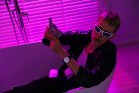 Young businessman in jacket and sunglasses with money in bath tub in violet light indoorsの写真素材