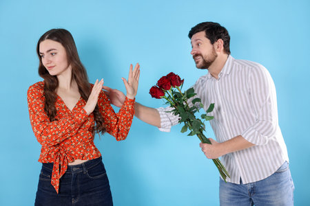 Embarrassed woman refusing bouquet of roses from man on light blue background