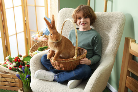 Little boy holding wicker basket with cute Easter bunny in armchair indoors