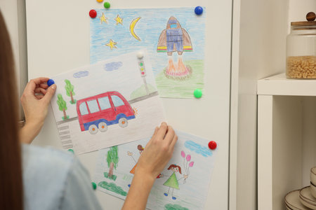 Woman attaching child's drawing of bus to white cabinet indoors, closeup