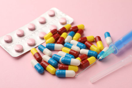 Different pills, blister and syringe on pink background, closeup